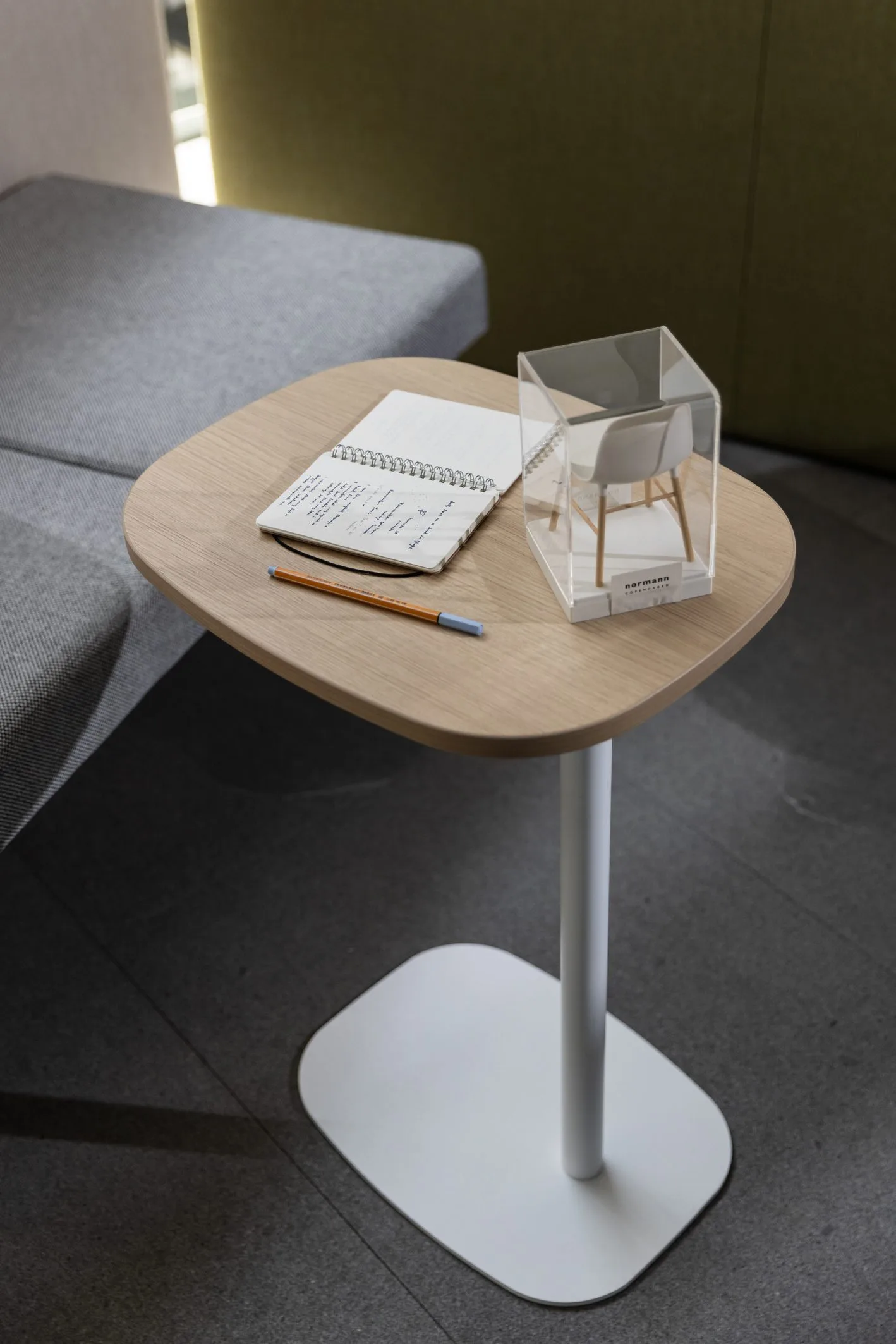Mesa auxiliar de madera clara con base blanca, junto a un sofá gris en el showroom de Poliarte