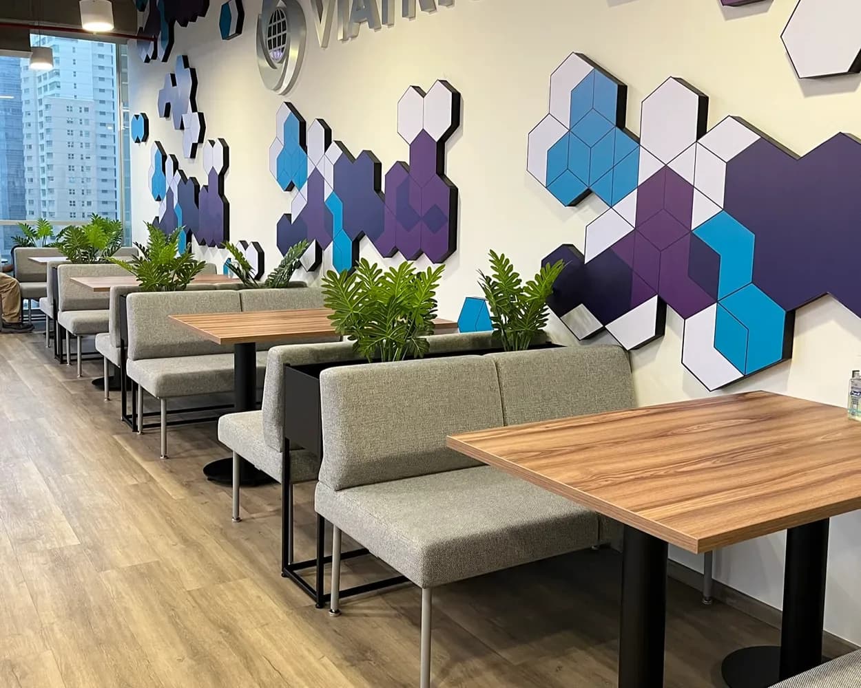 Comedor de Viatris con bancas tapizadas grises y mesas de madera junto a muro de hexágonos, por Poliarte