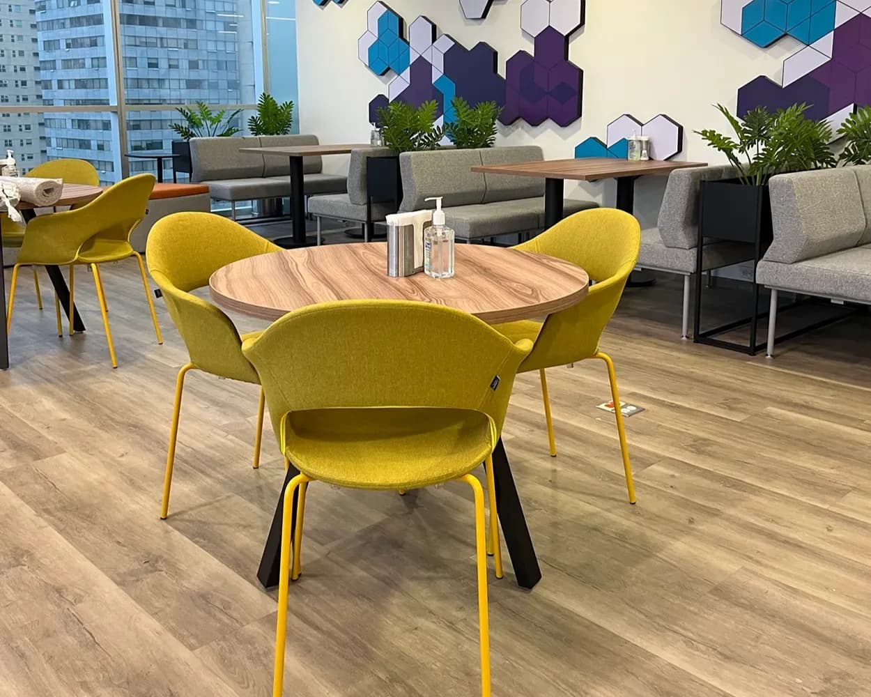 Mesa redonda de madera con sillas amarillas y bancas grises en el comedor de Viatris, por Poliarte